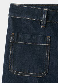 Poche arrière d'un jean en denim bleu foncé avec des coutures orange visibles et une boucle de ceinture sur un fond clair.