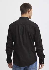 Camisa negra de manga larga con yugo trasero y detalle de pliegue, hecha de una tela texturizada. Cuenta con puños abotonados y un dobladillo curvado.