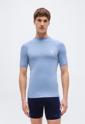 Νέος άνδρας που φοράει εφαρμοστή ανοιχτογάλαζια μπλούζα κολύμβησης και μαύρο σορτς κολύμβησης με μπλε λεπτομέρειες, στέκεται μπροστά σε απλό λευκό φόντο.