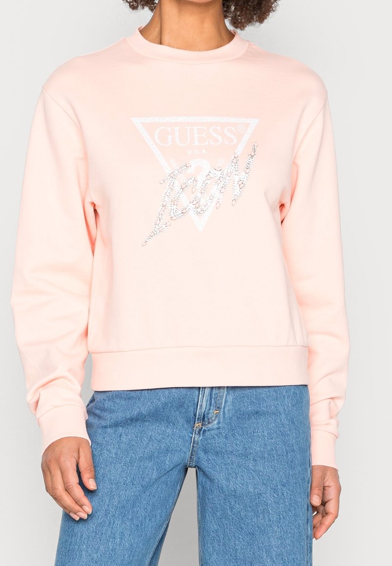 Personne portant un sweat-shirt Guess rose clair avec le texte "Icon" en strass sur un jean bleu, visible du cou aux hanches sur fond blanc.