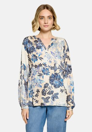 Kvinde med kort blondt hår iført en beige bluse med blå blomsterprint og blå jeans, stående mod en ensfarvet baggrund.