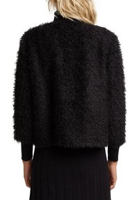 Svart fluffig jacka med en kortad design, texturerad yta och räfflade ärmar, som bärs över en svart ribbad klänning.