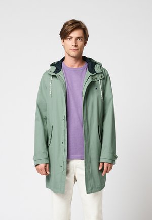 Jeune homme debout, portant une veste de pluie à capuche vert clair, un t-shirt violet et un pantalon blanc sur un fond uni.