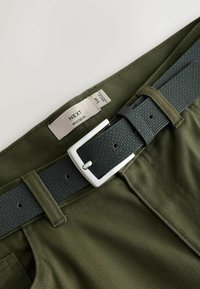 Zelené sportovní kalhoty s texturovaným tmavě šedým páskem se stříbrnou sponou. Obsahují štítek s názvem značky a velikostí.