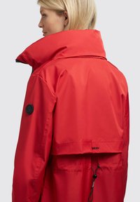 Chaqueta impermeable roja con un alto cuello, ajuste oversize y dobladillo ajustable. Presenta un parche de logotipo negro en la manga. Textura suave.