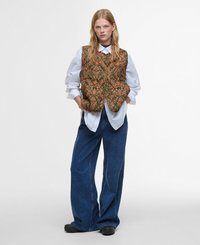Gequilted vest met een groen en oranje bloemenpatroon, gedragen over een wit shirt. Loose blauwe denim broek en zwarte hoge platform schoenen maken de look compleet.