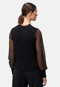 Schwarzer Pullover mit texturierten schwarzen Netzärmeln, die ein Fischgrätmuster aufweisen. Rundhalsausschnitt mit gewellten Kanten, taillierte Passform.