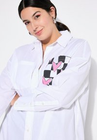 Camicia bianca con bottoni, caratterizzata da una tasca a scacchi bianca e nera con motivi a cuore rosa. Maniche lunghe con polsini a bottone. Tessuto di texture liscia.