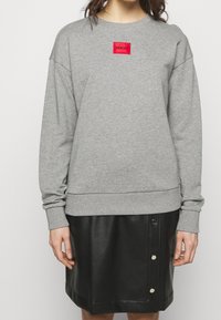 Osoba v šedém svetru s červenou nášivkou "HUGO" a černé kožené sukně na knoflíky, na jednobarevném světlém pozadí.