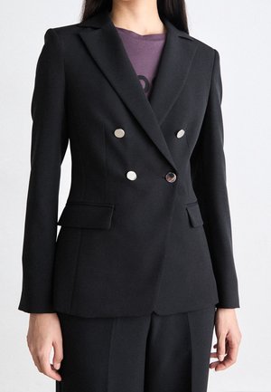 Žena v černém dvouřadém saku na zakázku se stříbrnými knoflíky, nošeném přes fialové tričko.