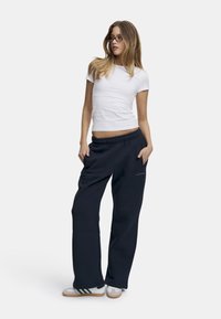 Hvit tettsittende t-skjorte kombinert med marineblå joggebukser med sidelommer. Modellen har på seg hvite sneakers med grønne striper og inntar en avslappet pose.