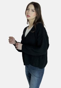 Schwarzer gestrickter Cardigan mit lockerer Passform, V-Ausschnitt und überschnittenen Schultern, kombiniert mit einem weißen Top und blauen Jeans. Sanfte Textur.