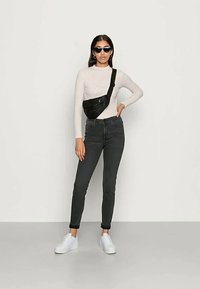 Mochila preta vestida sobre uma blusa de manga longa clara e jeans escuros, combinada com óculos de sol e tênis brancos, em pé sobre um fundo neutro.