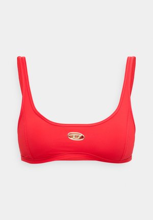 Roter Sport-BH mit dünnen Trägern und einem kleinen goldenen ovalen Logo, das mittig auf der Vorderseite vor einem schlichten weißen Hintergrund platziert ist.