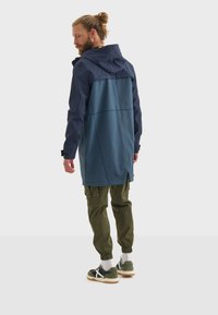 Homme portant une veste bicolore et un pantalon cargo olive, vu de derrière, debout sur un fond blanc. Chaussures décontractées visibles.