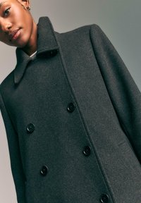 Giovane donna che indossa un cappotto di lana grigio scuro a doppiopetto con bottoni neri su uno sfondo neutro e semplice.