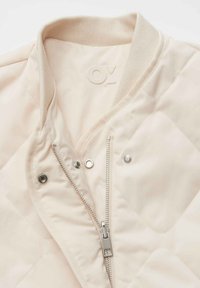 Veste matelassée beige clair avec col côtelé, fermeture éclair argentée partiellement ouverte et boutons-pression sur le devant.