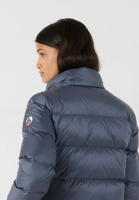 Giacca imbottita blu navy con colletto alto, caratterizzata da cuciture orizzontali e una patch con logo sulla spalla sinistra. Texture liscia e lucida.