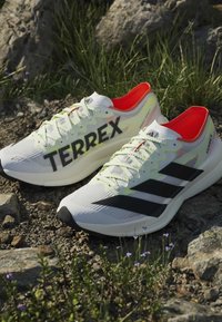 Ζευγάρι λευκά αθλητικά παπούτσια Adidas Terrex με μαύρες ρίγες και κίτρινα κορδόνια τοποθετημένα σε πετρώδες έδαφος με πράσινο γρασίδι.