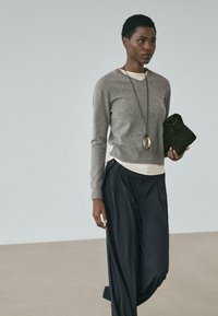 Grauer Wollpullover über einem weißen Oberteil, kombiniert mit lockeren schwarzen Hosen. Hält eine kleine dunkle Tasche und trägt eine lange silberne Anhängerkette.