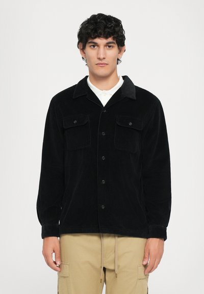 Giacca camicia in corduroy nera con colletto a punta, due tasche frontali e chiusura con bottoni. Indossata sopra una camicia bianca con maniche lunghe.