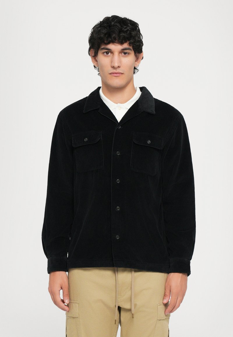 Black corduroy shirt jacket with a pointed collar, two front pockets, and button closure. Worn over a white shirt, featuring long sleeves.