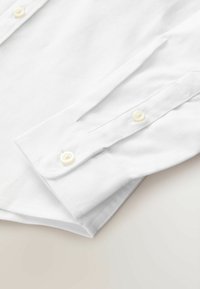 Weißes langärmeliges Hemd aus Baumwollstoff, mit einem Manschettenverschluss mit zwei gelblichen Knöpfen und einer dezenten Textur auf der Stoffoberfläche.