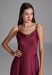 Robe bordeaux avec un décolleté en cowl et des bretelles fines. Fabriquée en tissu lisse, elle présente un corsage ajusté qui se déploie en une jupe ample.