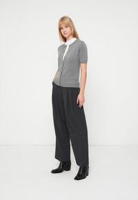 Siv kratki pulover s kratkimi rokavi s posameznim gumbom, nošen čez belo majico, kombiniran s črnimi širokimi hlačami z visokim pasom in črnimi gležnjači.