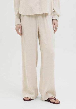 Haut beige, léger, ample et pantalon à jambes larges en tissu doux. Accentué de boutons et porté avec des tongs sombres.