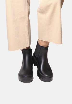 Zwarte enkellaarsregenlaarzen met grove zolen, gedragen met beige cropped broeken met rafelige zoom, tegen een effen achtergrond.