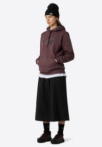 Burgunderhaze mit Logo, schwarzer Rock und dunkle Sneakers. Schichtweise mit einem weißen Shirt, schwarzem Beanie und markierten schwarzen Socken.
