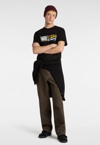 Svart grafisk t-shirt med "VANS"-logotyp, bruna baggybyxor och svarta sneakers. Tillbehören inkluderar en vinröd mössa och en svart tröja knuten runt midjan.