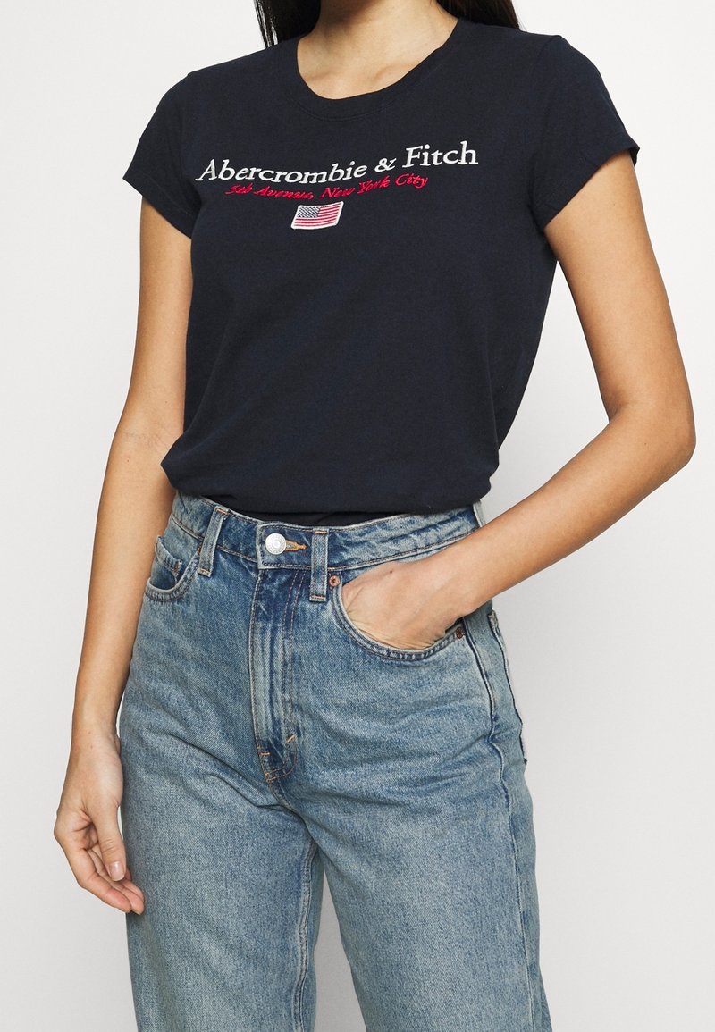 Camiseta de manga corta navy con texto blanco y rojo "Abercrombie & Fitch"; combinada con unos jeans de mezclilla de tiro alto en azul claro.