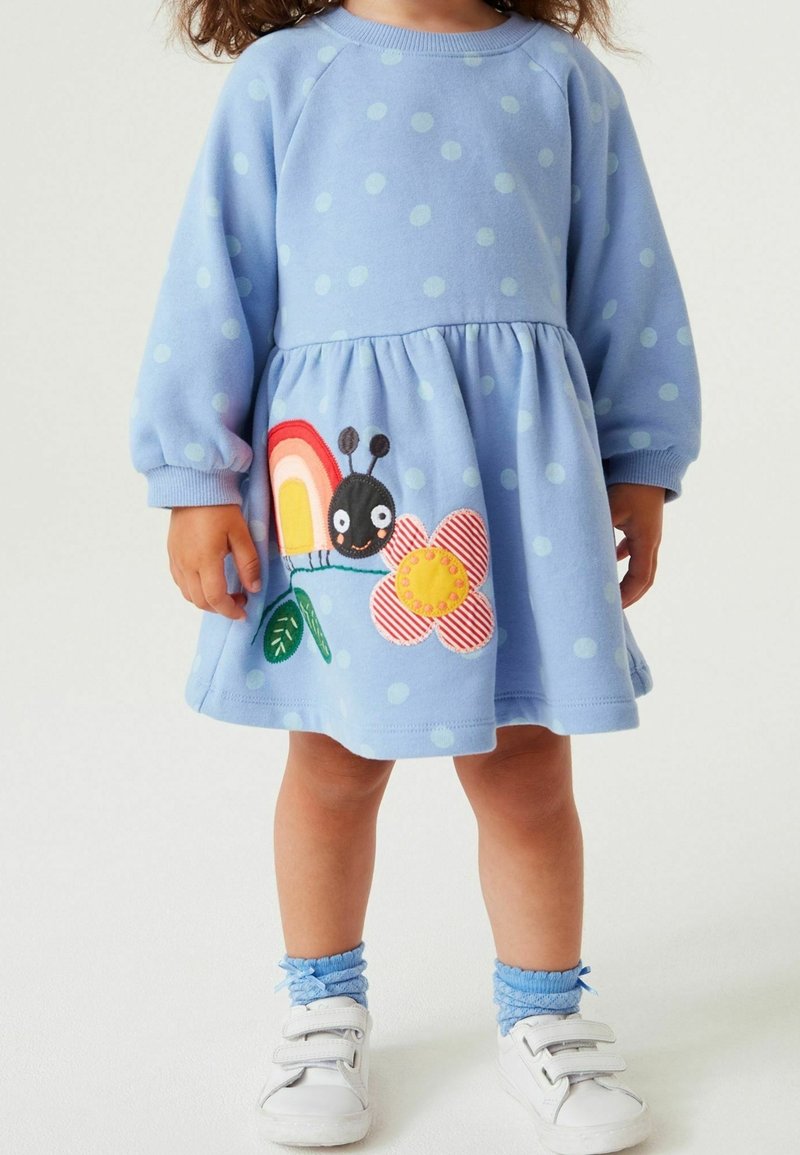 Tout-petit portant une robe bleu clair à pois avec une application colorée de chenille, fleur et arc-en-ciel sur la jupe, chaussettes bleues et chaussures blanches.