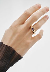 Anillo de oro con una gema circular de color púrpura, posicionado en una mano con una textura de piel ligeramente visible, contra un fondo claro.
