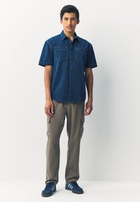 Chemise bleue à manches courtes avec un motif géométrique, boutonnée sur le devant, et deux poches poitrine. Associée à un pantalon cargo beige et des baskets bleues.