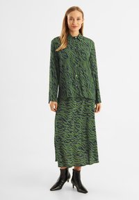 Abito verde con motivo ondulato e bottoni, abbinato a stivaletti neri alla caviglia. Maniche lunghe e design della gonna fluida.