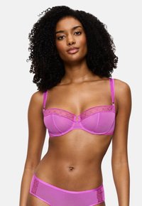 Sieviete ar cirtainiem matiem, kas valkā atbilstošu spilgti rozā caurspīdīgu krūšturi un biksītes, stāv uz neitrāla gaiša fona.