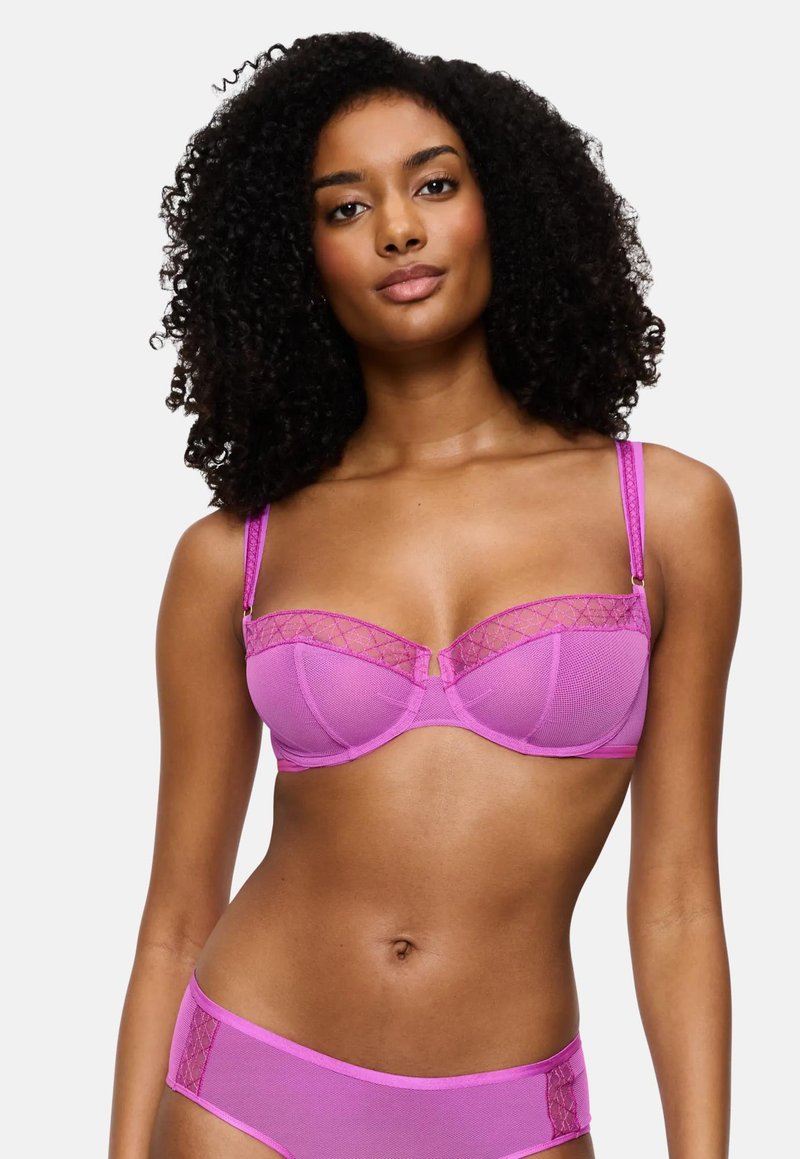 Sieviete ar cirtainiem matiem, kas valkā atbilstošu spilgti rozā caurspīdīgu krūšturi un biksītes, stāv uz neitrāla gaiša fona.