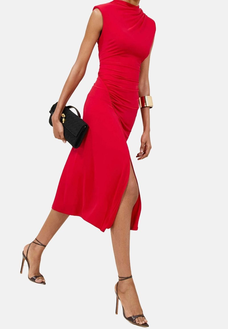 Femme marchant dans une robe rouge sans manches avec une fente latérale, des talons hauts noirs, une pochette noire et un bracelet manchette en or au bras.