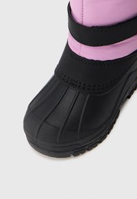 Černá gumová bota s texturovanou podrážkou a růžovou nylonovou horní částí, jištěná černými textilními popruhy. Obsahuje zakřivenou špičku a vroubkované detaily.