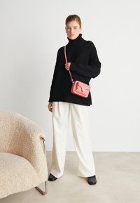 Maglione nero a coste con collo alto, pantaloni bianchi a gamba larga, borsa a tracolla rosa con finiture dorate, scarpe nere lucide, poltrona beige testurizzata.