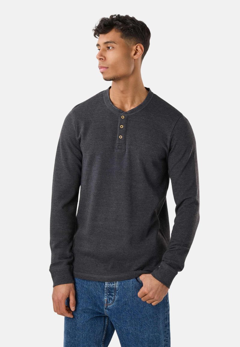 Mann trägt dunkelgraues Henley-Langarmshirt mit drei Knöpfen, blaue Jeans, schaut nach links, Hand in der Tasche.
