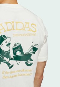 Bílá bavlněná tričko s zeleno-žlutým grafickým potiskem muže s kolečkem a golfovými holemi, s nápisem "ADIDAS GROUNDSKEEPING".
