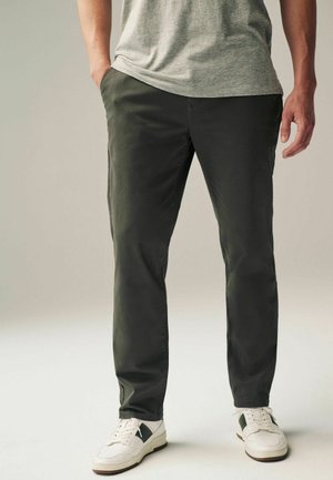 Pantalon slim-fit vert foncé en coton doux. Présente des poches latérales et un design à plat à l'avant. Associé à des baskets blanches et une chemise grise.