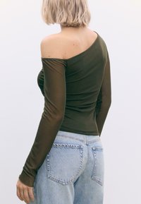 Olivengrøn, top med én skulder og lange, tætsiddende ærmer; glat tekstur, parret med lyseblå højtaljede jeans med baglommer.