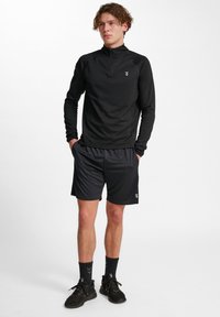 Zwarte, lange-mouw sporttop met een ritskraag, gecombineerd met zwarte shorts. Heeft een subtiele logo op zowel het shirt als de shorts.