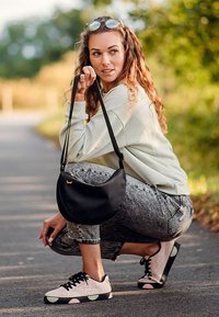 Černá krosová taška s zakřiveným designem, kterou má na sobě osoba ve světle šedém svetr a vzorovaných džínách, s černými a světle růžovými teniskami.