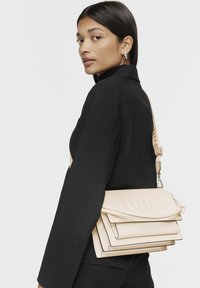 Borsa a tracolla in pelle beige con logo "TOUS" in rilievo, design a strati, tracolla regolabile e superficie liscia. Modella che indossa un outfit nero.